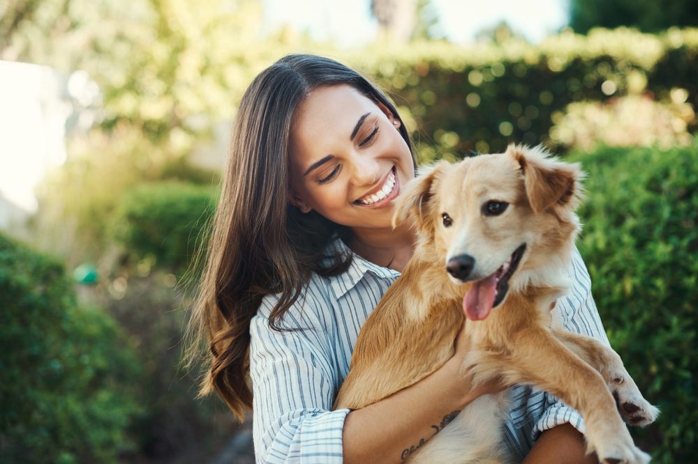 A smiling young woman with long brown hair, wearing a white and blue striped shirt, holding a golden-colored, medium-sized dog with its tongue out in her arms in an outdoor, leafy garden setting on a sunny day. A dog loves Superfood Science Plant-based protein all natural dog treats. 